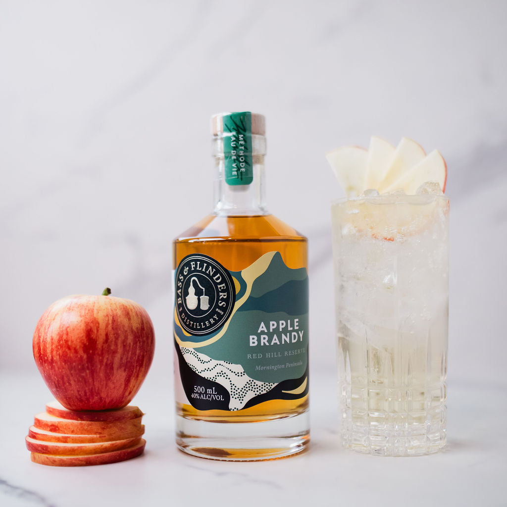 Bottle of Bass & Flinders Distillery Apple Brandy with an apple and a tall glass of apple brandy spritz cocktail on a white background