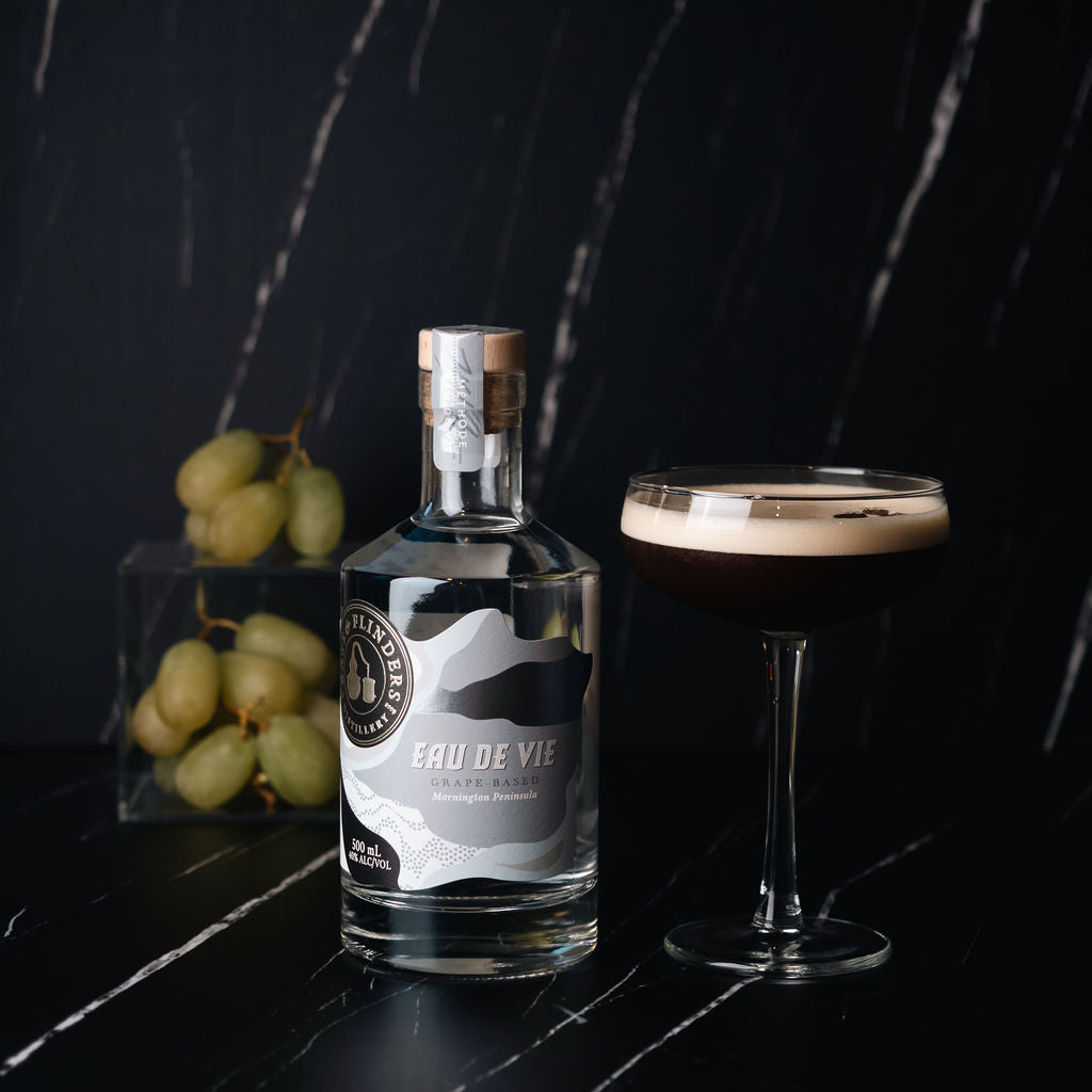 Bottle of Bass & Flinders Distillery Eau de Vie spirit on a dark surface with grapes and a coupe cocktail glass containing an eau de vie espresso martini cocktail.