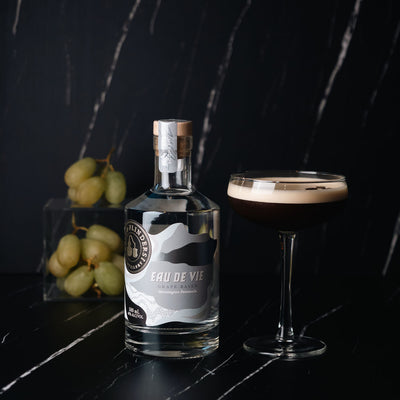 Bottle of Bass & Flinders Distillery Eau de Vie spirit on a dark surface with grapes and a coupe cocktail glass containing an eau de vie espresso martini cocktail.