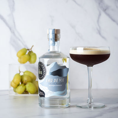 Bottle of Bass & Flinders Distillery Eau De Vie spirit with a coupe cocktail glass containing a layered espresso martini cocktail on a white surface with grapes in the background.