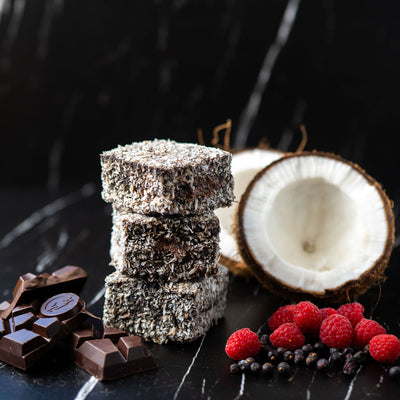 Bass & flinders distillery lamington gin ingredients including Stacks of chocolate and lamingtons with half a coconut, raspberries and juniper berries on a dark marble surface