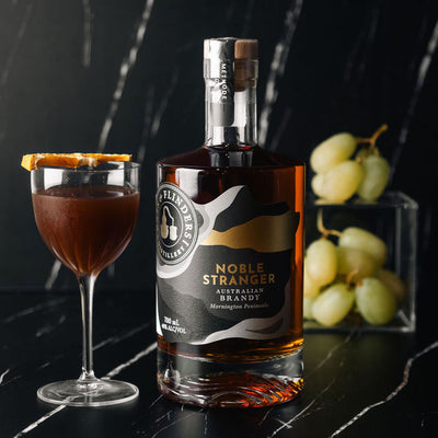 Bottle of Bass & Flinders Distillery Noble Stranger brandy with a cafe sazerac brandy cocktail in glass and bunch of grapes on a dark surface